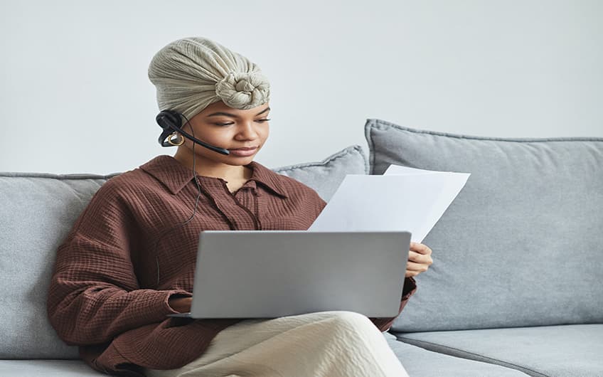 Black Woman working with her laptop computer while using a headset to talk to a client